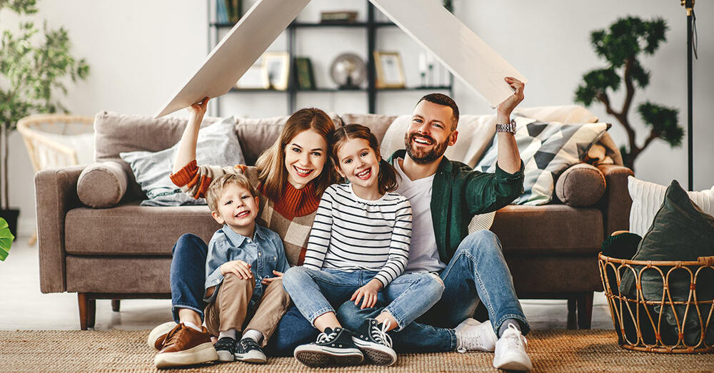 Familie mit zwei Kindern sitzt lachend im Wohnzimmer vor der Couch.