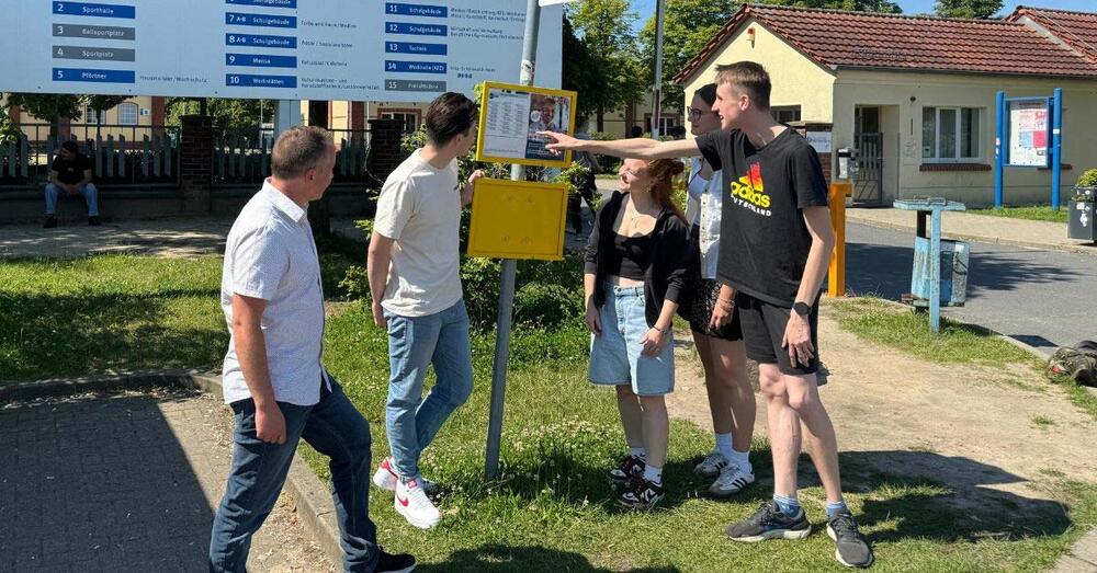Sch&uuml;ler stehen an einer Bushaltestelle und schauen sich den Busfahrplan an