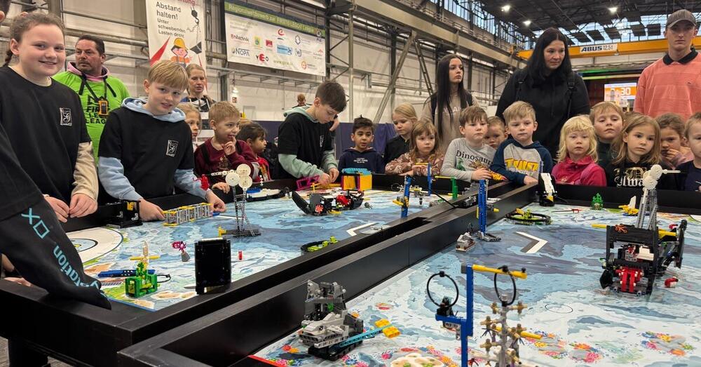 Sch&uuml;ler stehen mit Lehreren in der Trainingshalle des Qualifizierungscentrum der Wirtschaft um zwei Tische herum, auf den verschiedene komplexe Lego-Konstruktionen aufgebaut wurden.